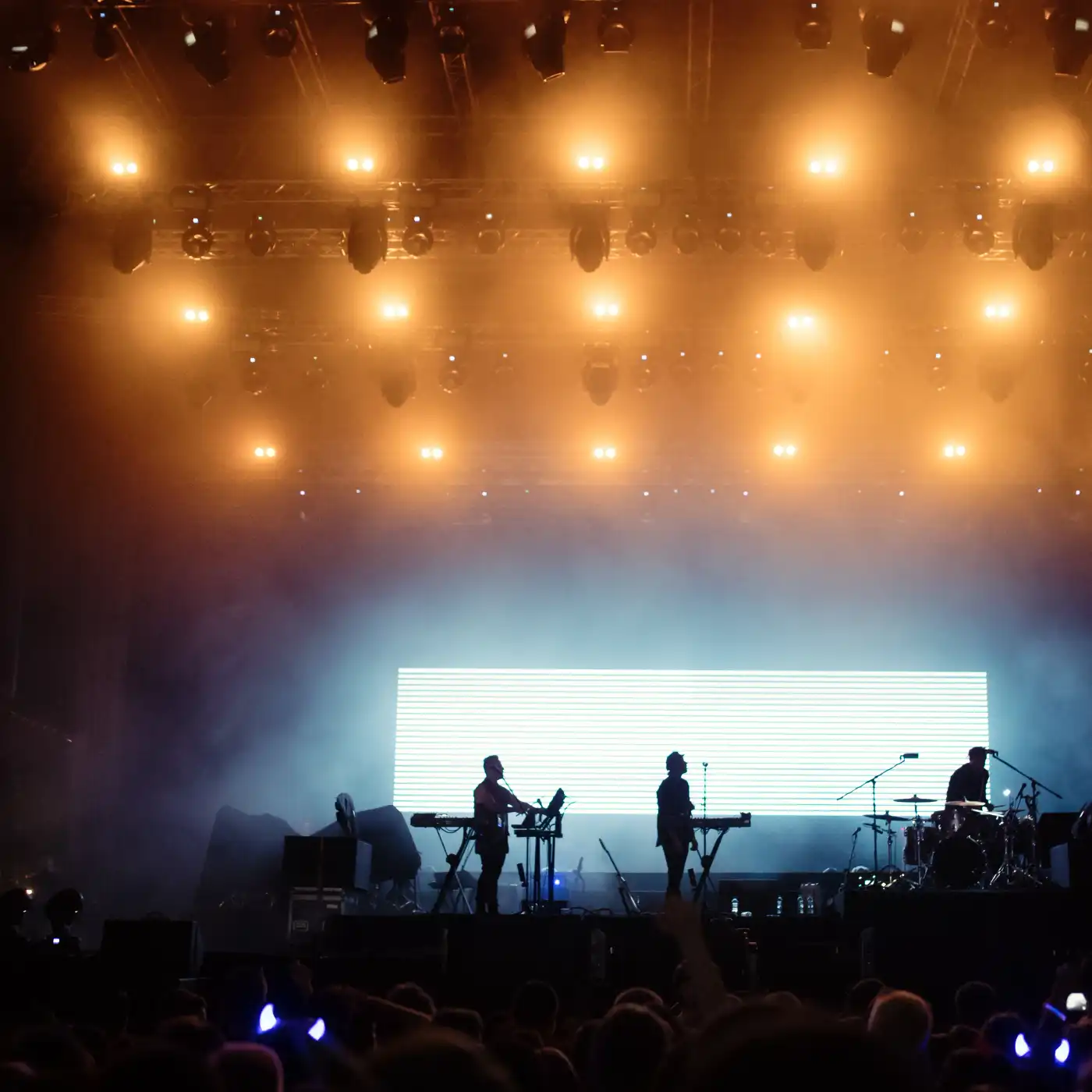 Image of a concert hall with big bright orange lights on the ceiling and on stage playing a three man band.