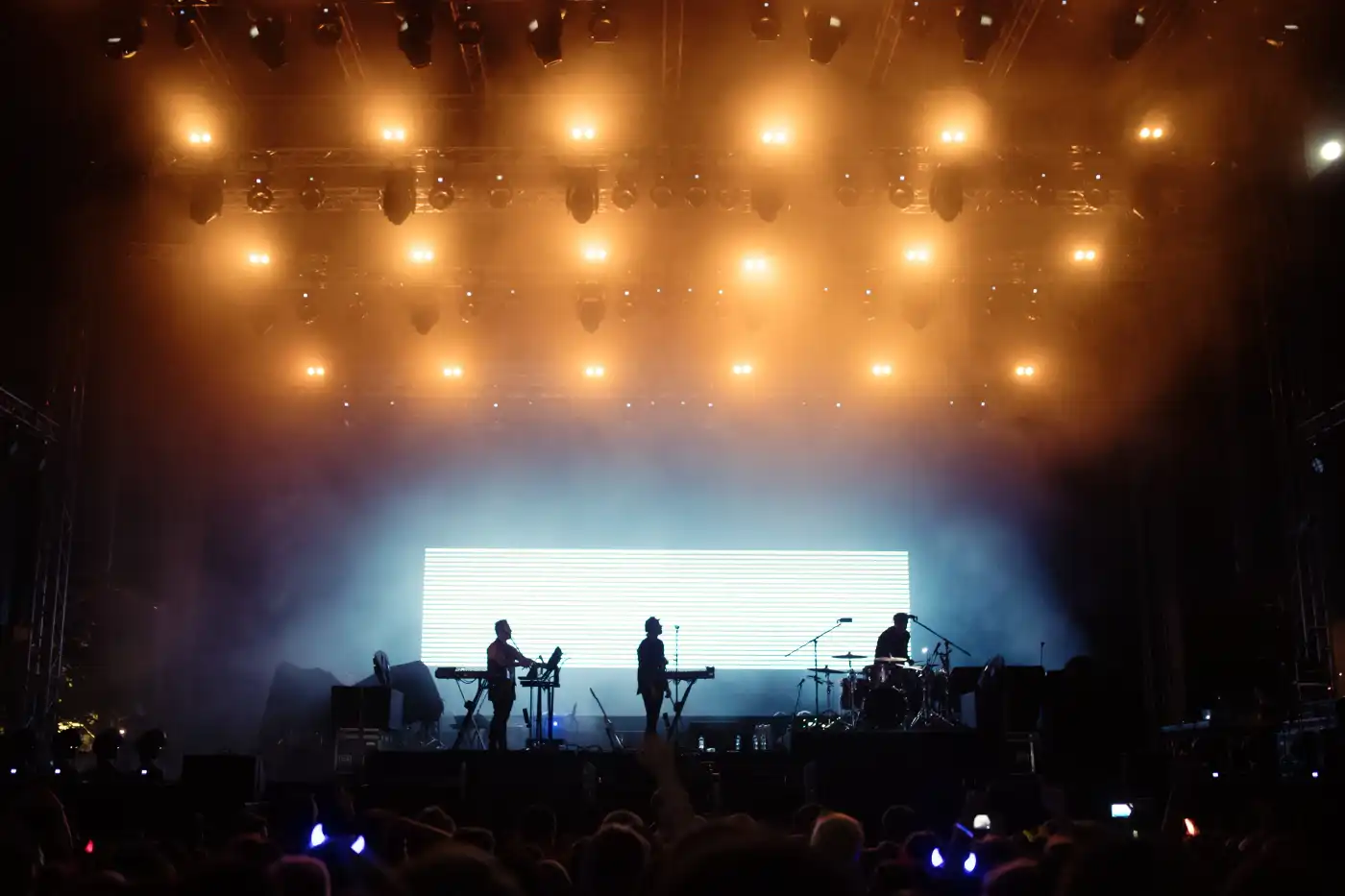 Image of a concert hall with big bright orange lights on the ceiling and on stage playing a three man band.